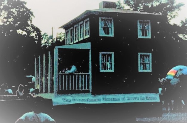 A replica of the 2 story Lock House with people on the porch on a float for a Havre de Grace parade