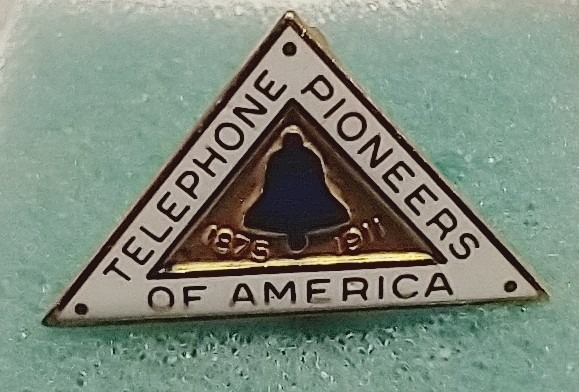 A triangular-shaped pin that reads: Telephone Pioneers of America, 1975-1911, and has an image of the Bell system's traditional bell.